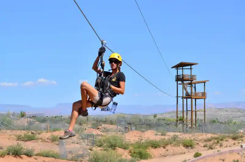 A rider in a harness and helmet flying along a zip line high above the Camp Verde landscape